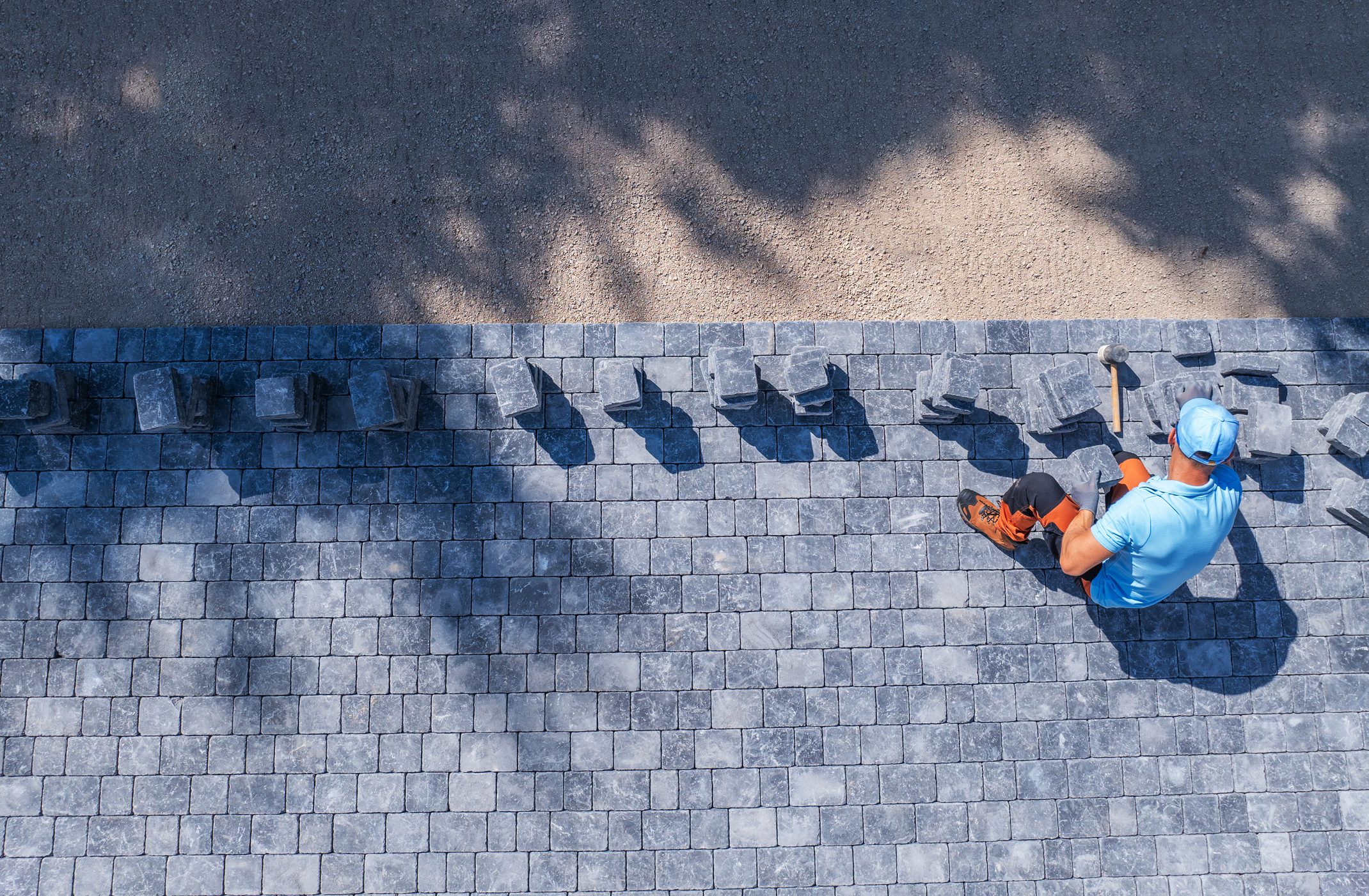 A construction worker carefully positions pavers on a pathway, surrounded by tools and materials in bright sunlight.